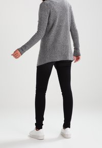 Person wearing a grey herringbone-patterned sweater, black slim pants, and white sneakers, standing against a plain background.