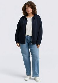 Felpa con cappuccio blu navy con cerniera e coulisse, indossata sopra una camicia bianca. Abbinata a jeans azzurri e sneakers bianche. Tessuto liscio, vestibilità comoda.