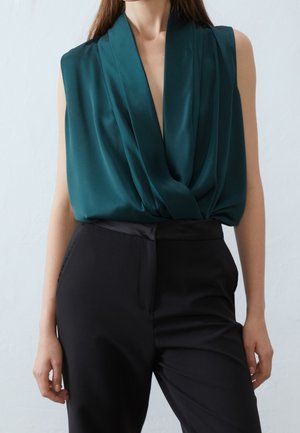 Blusa sin mangas drapeada en verde oscuro metida dentro de pantalones negros de talle alto y corte sastre, vista en una persona de pie.