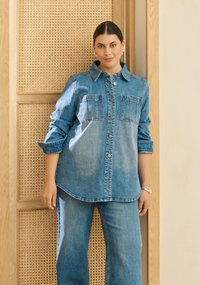 Chemise en jean boutonnée avec col classique, dotée de deux poches poitrine, manches retroussées et ourlet légèrement arrondi, assortie à un jean de la même matière.