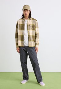 Chemise en flanelle à carreaux olive et beige avec une poche poitrine, pantalon gris et baskets lilas claires. T-shirt blanc uni en dessous. Fond vert.