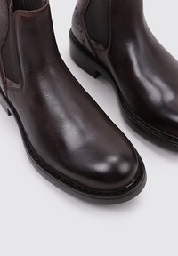 Bottines Chelsea en cuir marron avec une texture lisse, bout arrondi, panneaux latéraux élastiques et une semelle noire robuste. Détails de couture présents.