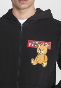 Crna dukserica sa kapuljačom i patentnim zatvaračem, s grafikom žutog medvjedića i crvenim natpisom "KAVANACH" na prsima. Mekana tkanina s ležernim krojem.