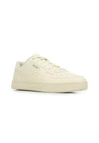 Sneaker Puma a basso profilo di colore crema con design lacciato, punta perforata e striscia laterale con il logo su sfondo bianco.