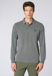 Polo shirt grigio a maniche lunghe con colletto, dotato di un'emblema nera sul petto, abbinato a pantaloni verde oliva. Tessuto morbido e testurizzato.
