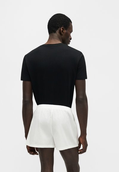 Muž viditeľný zozadu, oblečený v jednofarebnom čiernom tričku s krátkym rukávom a voľných bielych krátkych nohaviciach na jednofarebnom svetlom pozadí.