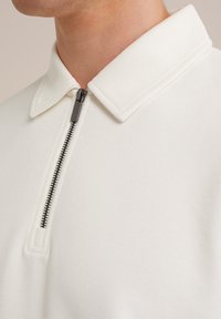 Polo blanco con cremallera y tejido suave y texturizado, que presenta un collar puntiagudo y un detalle de cremallera negra en contraste.