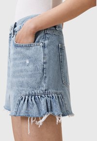 Jupe en jean bleu clair avec ourlet effiloché et détail volanté, portée par une personne avec la main dans la poche avant sur un fond uni.