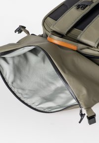 Zaino verde oliva con un esterno liscio e resistente e un interno grigio chiaro. Include cerniere nere e un accentuazione in pelle marrone.