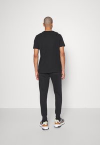 Svart bomull T-shirt med korta ärmar, tillsammans med svarta sweatpants och flerfärgade sneakers, sedd bakifrån mot en enfärgad bakgrund.