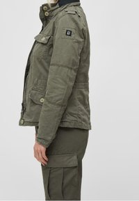 Olivgrön militärinspirerad jacka med knäppning fram, flera fickor och ett svart logopatch på vänster ärm. Strukturerad tyg.