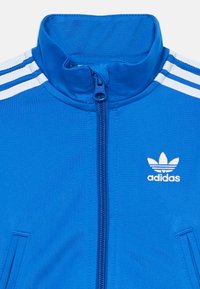 Blå Adidas träningsjacka med vita ränder på axlarna, full dragkedja fram och vit Adidas trefoil-logotyp på bröstet.