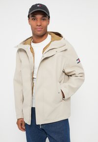 Hombre con chaqueta con capucha beige sobre una capa con cremallera color beige claro y camisa blanca, con gorra gris oscuro y jeans azules, de pie contra un fondo liso.
