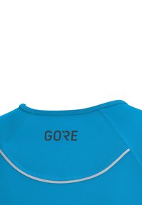 Ein türkisfarbenes Sportshirt mit rundem Ausschnitt, reflektierenden Paspeln und einem auffälligen schwarzen Logo "GORE" auf dem Rücken.