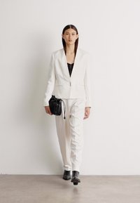 Blazer branco ajustado e calças combinando, acompanhados por um top preto cropped e uma pequena bolsa preta. Tecido suave, design minimalista, forma ajustada.
