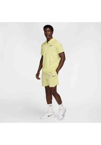 Poloshirt und Shorts-Set aus leichtem Polyester in Hellgelb, mit einem schwarzen Nike-Swoosh-Logo, kombiniert mit weißen Sportschuhen und Socken.