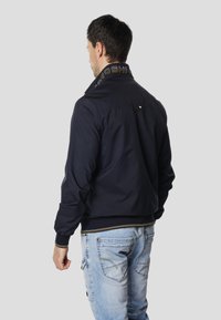 Námořnicky modrá bomber bunda s vyšívaným límcem, zapínáním na zip a elastickými manžetami. Nošená přes světle modré džíny, které ukazují uvolněný střih.