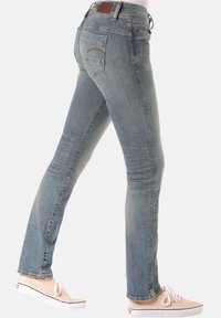 Slim-fit blå jeans med bakfickor som är sydda, bärs med ljusrosa canvas-sneakers på en person som går.