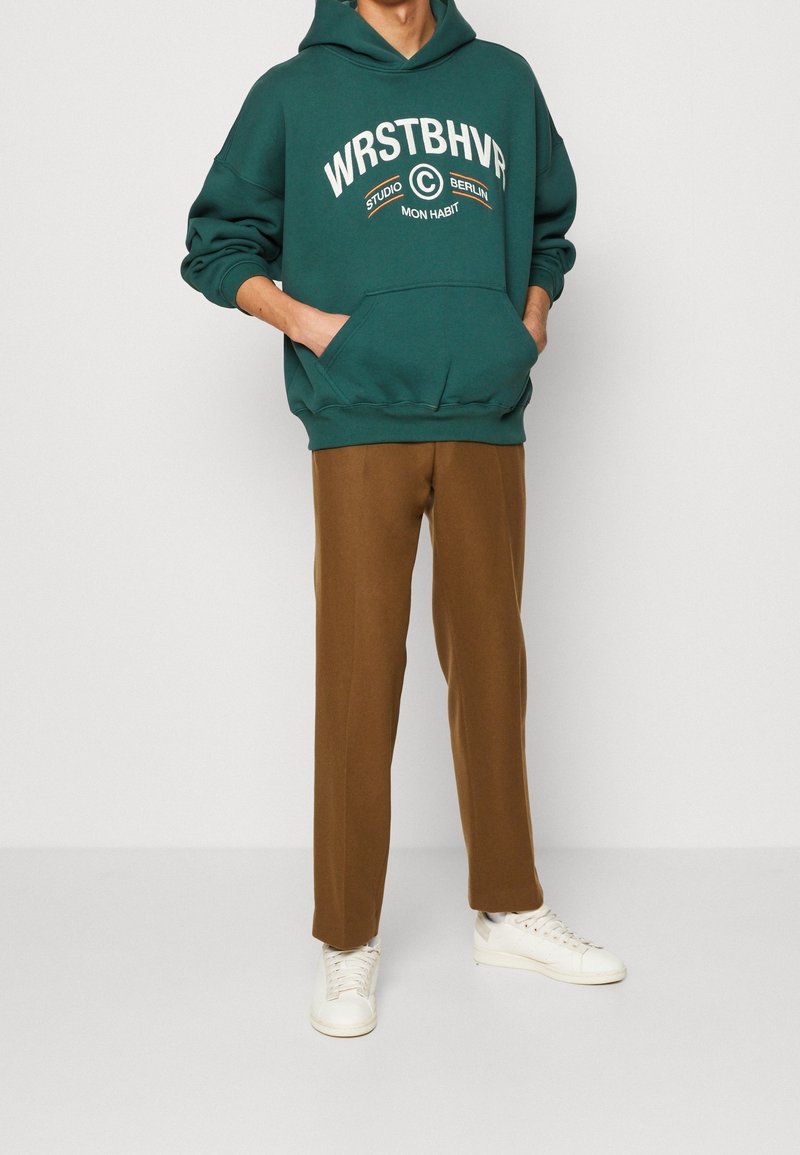 Personne portant un sweat à capuche vert foncé avec le texte "WRSTBHVR", un pantalon marron droit et des baskets blanches, debout avec les mains dans les poches du sweat.