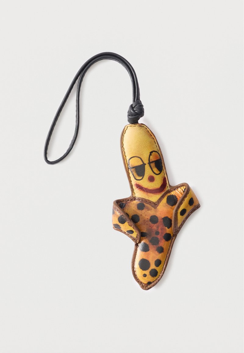 Μπρελόκ από δέρμα σε σχήμα ξεφλουδισμένης μπανάνας με μαύρα στίγματα και ζωγραφισμένο χαμογελαστό πρόσωπο, προσαρτημένο σε μαύρο κορδόνι.