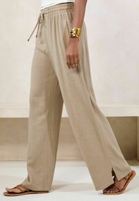 Pantaloni beige a gamba larga con vita regolabile, tasche laterali e leggeri spacchi all'orlo, abbinati a sandali piatti marroni.