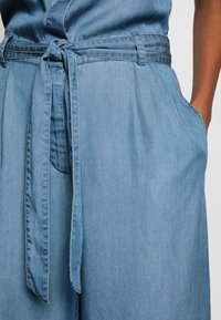 Tutina in denim blu chiaro con vita regolabile, caratterizzata da un davanti annodato e tasche laterali. Il tessuto ha una consistenza liscia e una vestibilità comoda.