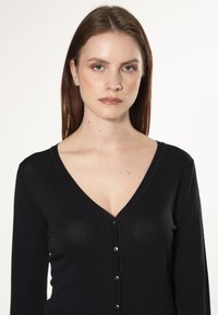 Cardigan nero con maniche lunghe e scollatura a V. Presenta una chiusura con bottoni sul davanti, un leggero lucido e una silhouette aderente.