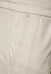 Gros plan sur un pantalon à taille élastique beige montrant une poche latérale avec des coutures visibles et un petit rivet métallique.