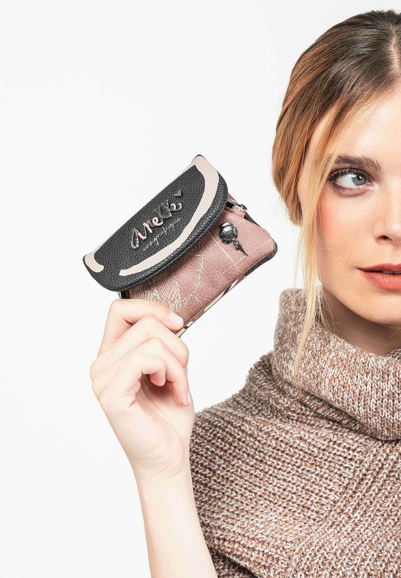 Mujer vestida con un suéter de punto beige sostiene un pequeño monedero negro y rosa con un broche de metal y un encanto decorativo.