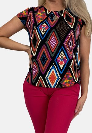 Femme portant un haut à manches courtes avec des motifs géométriques colorés en forme de losange et un pantalon rose vif, la main posée sur la hanche.