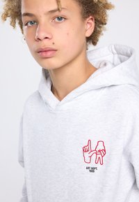 Grijze hoodie met een voorvak, met een rood geborduurd ontwerp van handen en de tekst "ART DEPT. 1989." Zachte, gestructureerde stof.