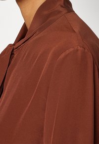 Blouse en satin marron avec un col noué et une texture douce et lisse, coupe décontractée avec détails froncés aux manches.