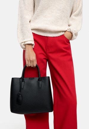 Osoba ubrana w kremowy sweter i czerwone spodnie trzyma czarną torebkę ze skóry o fakturze w jednej ręce za uchwyty.