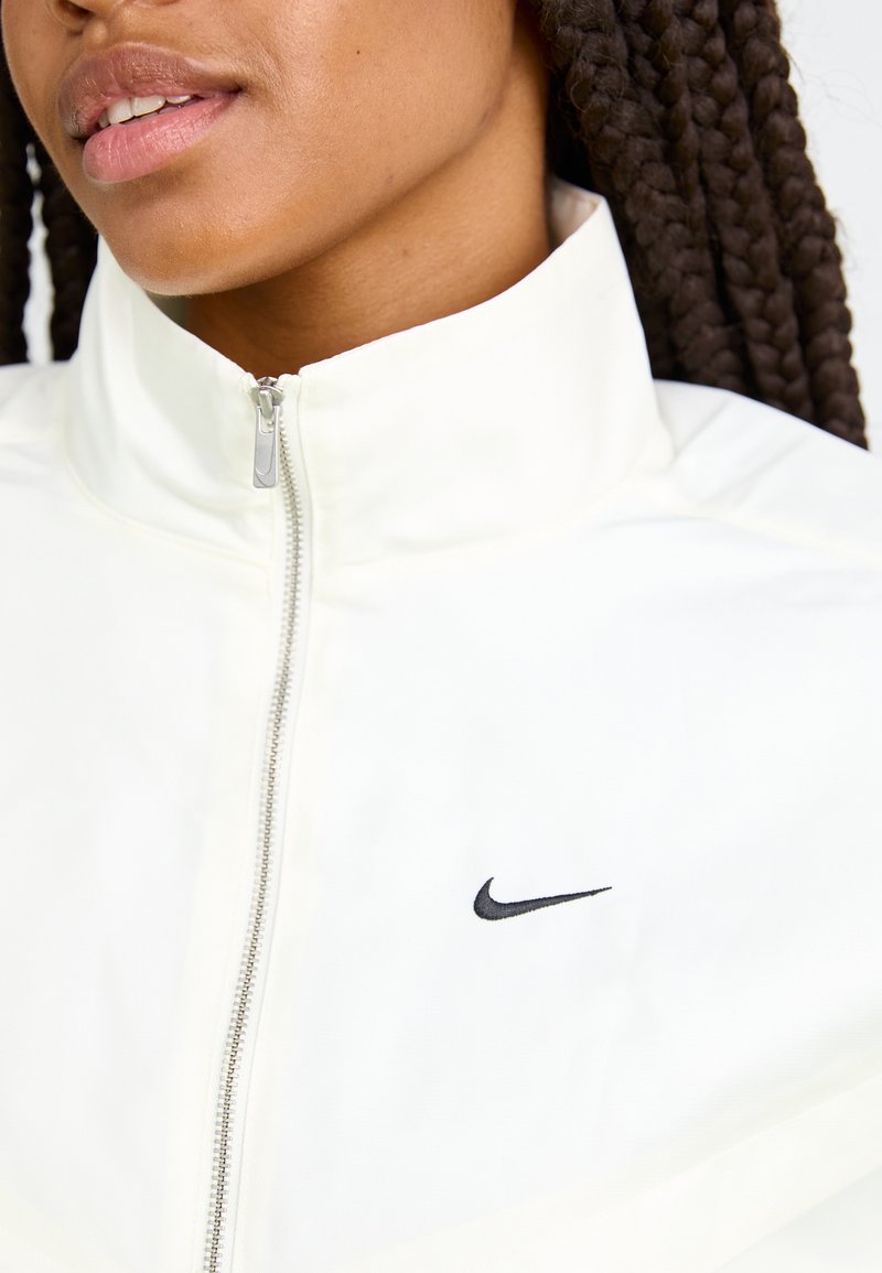 Close-up of a person wearing a white zip-up jacket with a black Nike swoosh logo on the chest and braided hair visible.