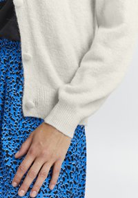 Cárdigan blanco de punto con botones redondos y puños acanalados, combinado con una falda azul decorada con un patrón de print de leopardo negro.