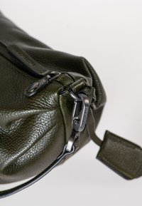 Borsa in pelle verde scuro con finitura testurizzata, dotata di chiusura con zip e accessori metallici neri. Include una tracolla rimovibile e un'etichetta decorativa.