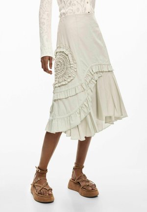 Femme portant une jupe midi crème à volants avec un détail plissé circulaire, associée à des sandales plateformes marron clair avec brides à la cheville.