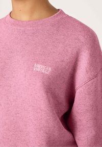 Roza svetrovka iz mehkega blaga z teksturiranim zaključkom. Ima okroglo ovratnik in vezen logotip "AMERICAN VINTAGE" na prsih.