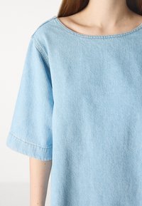 Haut en denim bleu clair avec des manches courtes et un col rond large. Le tissu a une texture douce avec des détails de surpiqûre subtils.