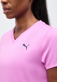 Κοντινό πλάνο ενός ατόμου που φορά ένα ανοιχτό ροζ μπλουζάκι Puma με V-λαιμόκοψη και μαύρο λογότυπο Puma στο αριστερό στήθος.