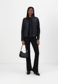 Vesta puf gri închis cu buzunare cu fermoar, purtată peste un hanorac negru. Pantaloni negri evazați și cizme cu toc negre elegante. Geantă mică de mână neagră.