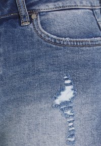 Blå denimjeans med en sliten reva på framsidan av fickan, med kontrasterande gul sömnad och en jämn yta.