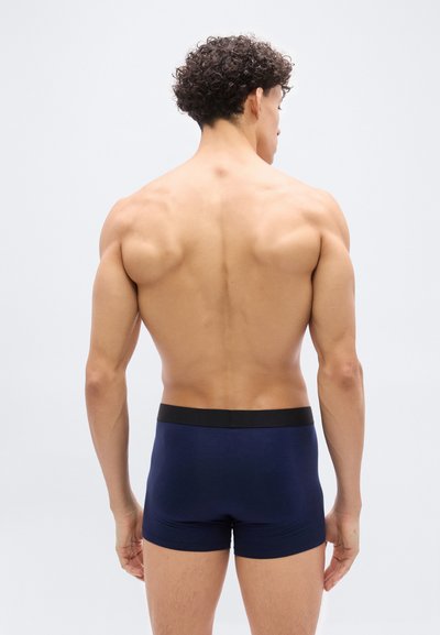 Άνδρας με σγουρά μαλλιά στέκεται με την πλάτη, φοράει σκούρο μπλε μποξεράκια με μαύρη μέση, μπροστά σε ένα απλό φωτεινό φόντο.