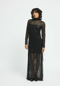 Rochie neagră cu mâneci lungi, realizată din material transparent, cu detalii de franjuri și o siluetă ajustată, care se extinde până la podea.