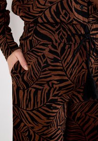 Brown fabric with black leaf pattern, featuring a drawstring waist and a side pocket. The texture appears soft with a slight sheen.