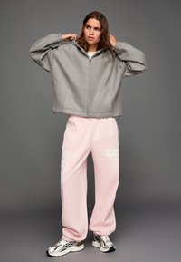 Šedá, nadmerná bunda na zips s jemnou textúrou a kapucňou. Kombinovaná s ružovými teplákmi s bielym textom a striebornými teniskami.