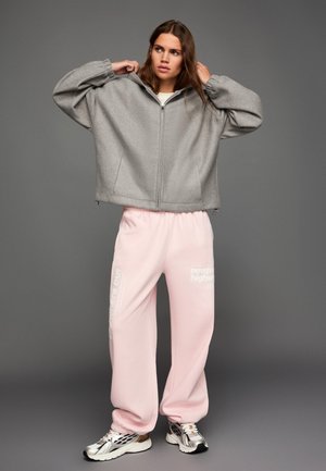 Šedá, nadmerná bunda na zips s jemnou textúrou a kapucňou. Kombinovaná s ružovými teplákmi s bielym textom a striebornými teniskami.