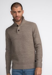 Pulover de culoare maro deschis, tricotat texturat, cu guler înalt și două butoane la gât. Mâneci și tiv cu model raiați. Purtați deasupra unor blugi din denim.