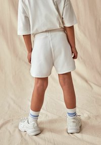 Shorts en coton blanc avec taille élastique, associés à un t-shirt blanc ample, des baskets blanches et des chaussettes rayées blanches, vus de derrière.