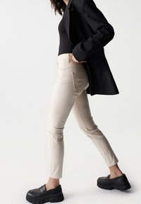 Pantalones ajustados color beige, hechos de denim con una textura sutil. Cuentan con bolsillos traseros con un detalle acanalado. Combinados con zapatos negros de estilo chunky sin cordones.